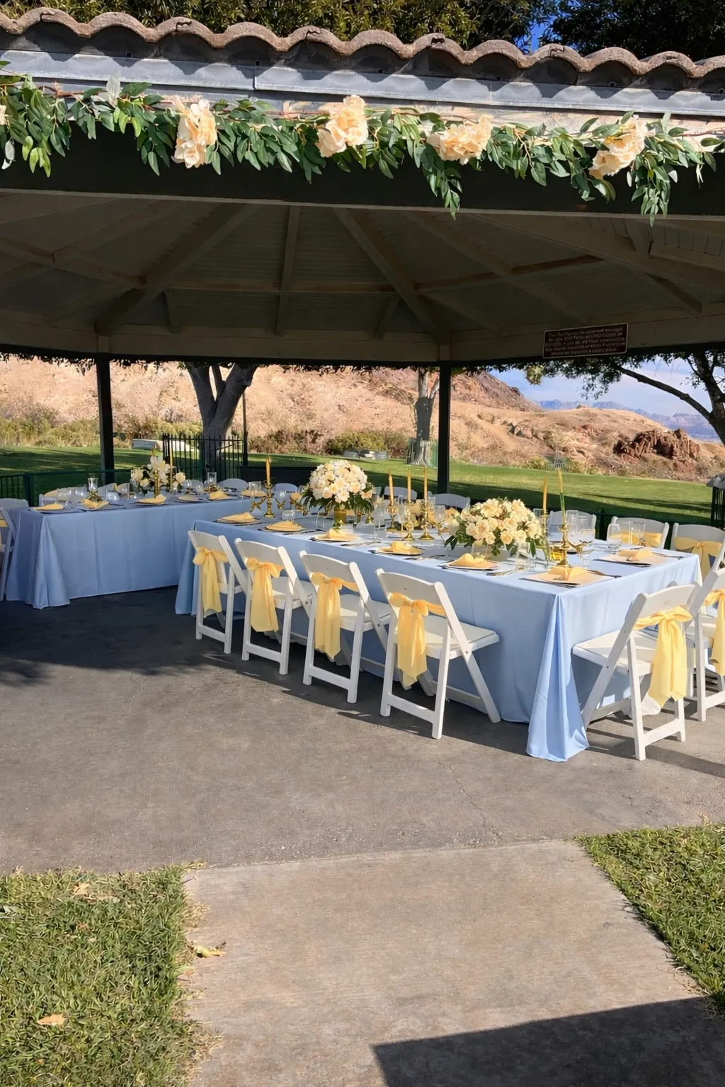 Elegant outdoor pavilion wedding setup with soft blue linens, butter yellow chair sashes, and floral centerpieces by Joyful Moments Party Rentals