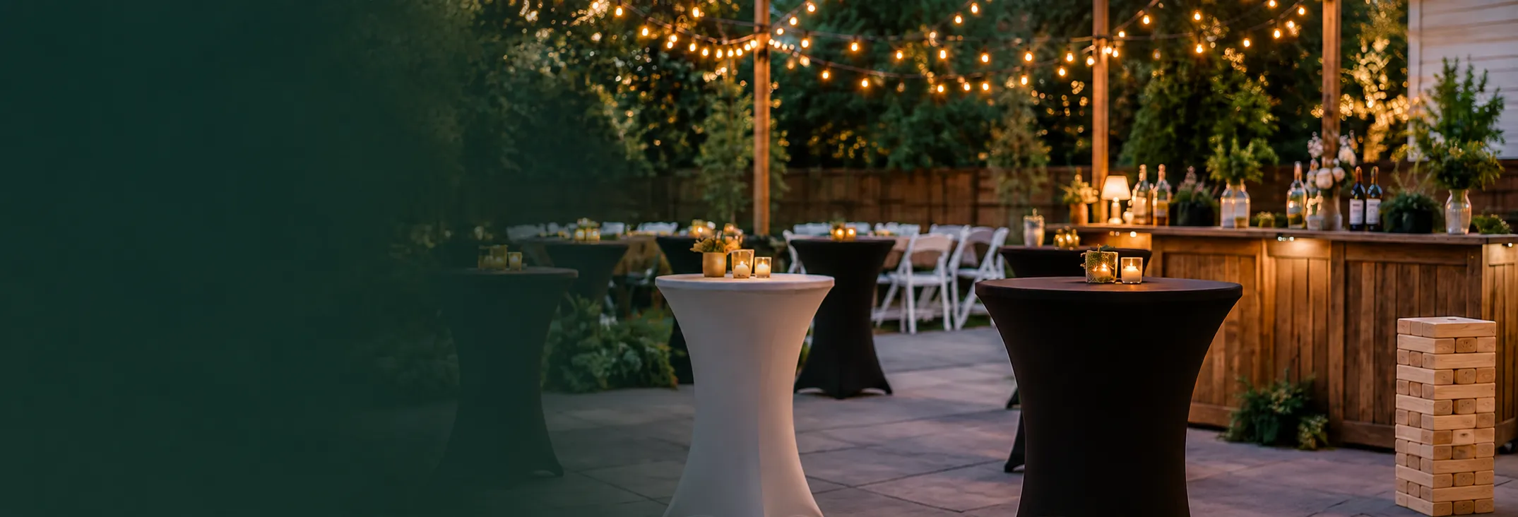 Outdoor party setup with cocktail tables, bar, white chairs and oversized wood block game under string lights