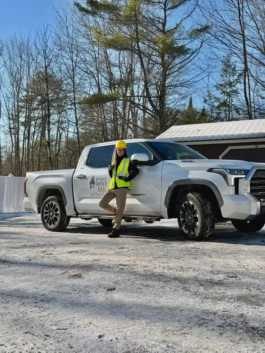 Becca, the owner and operator of Event Rent ME poses in front of her 2025 Toyota Tundra Limited iForce Max branded with the logo. Becca is wearing a hardhat, gloves, a bright reflective vest, and steel toe boots.