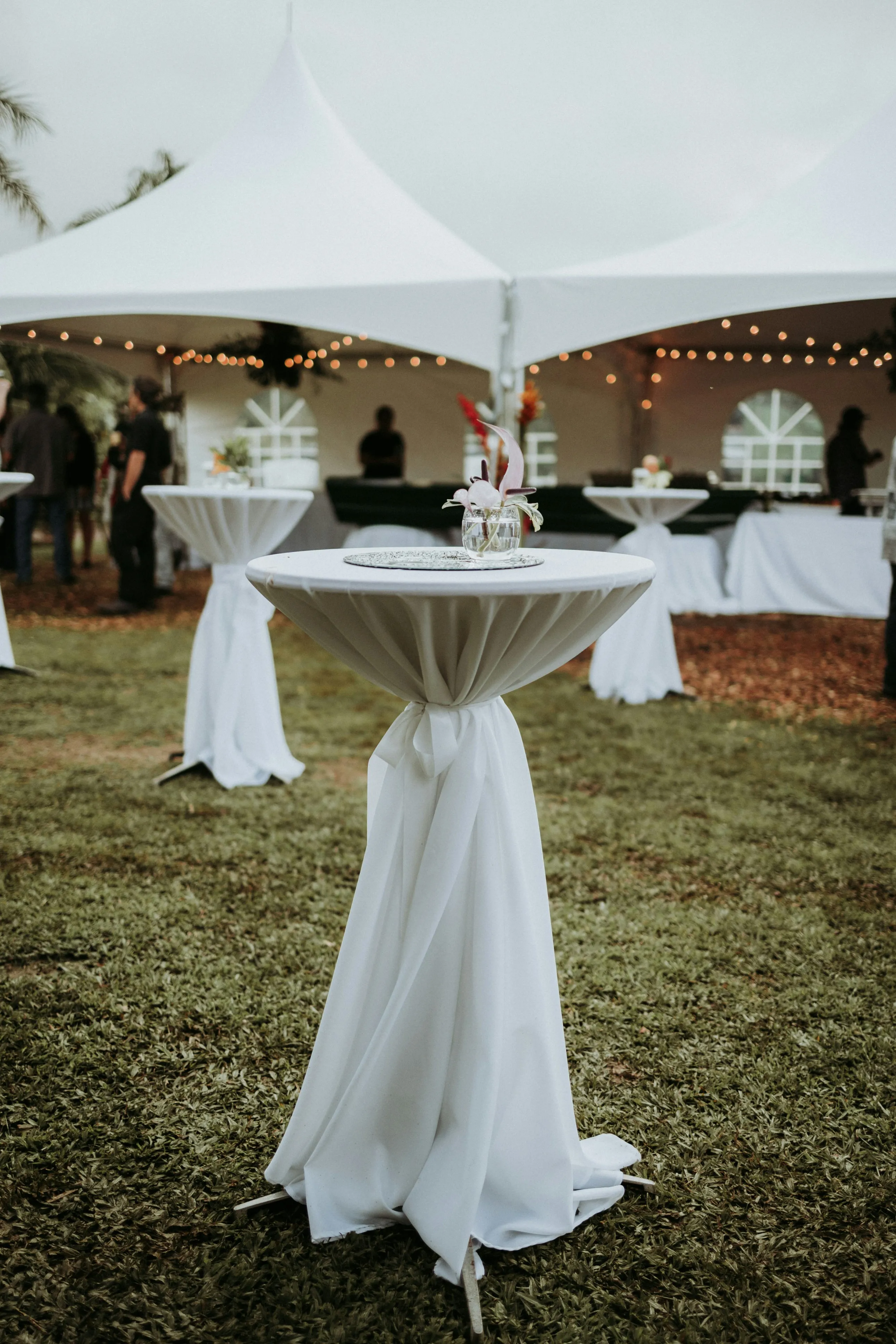 Standing tables draped with elegant tablecloths with a high peak frame tent in the background. 