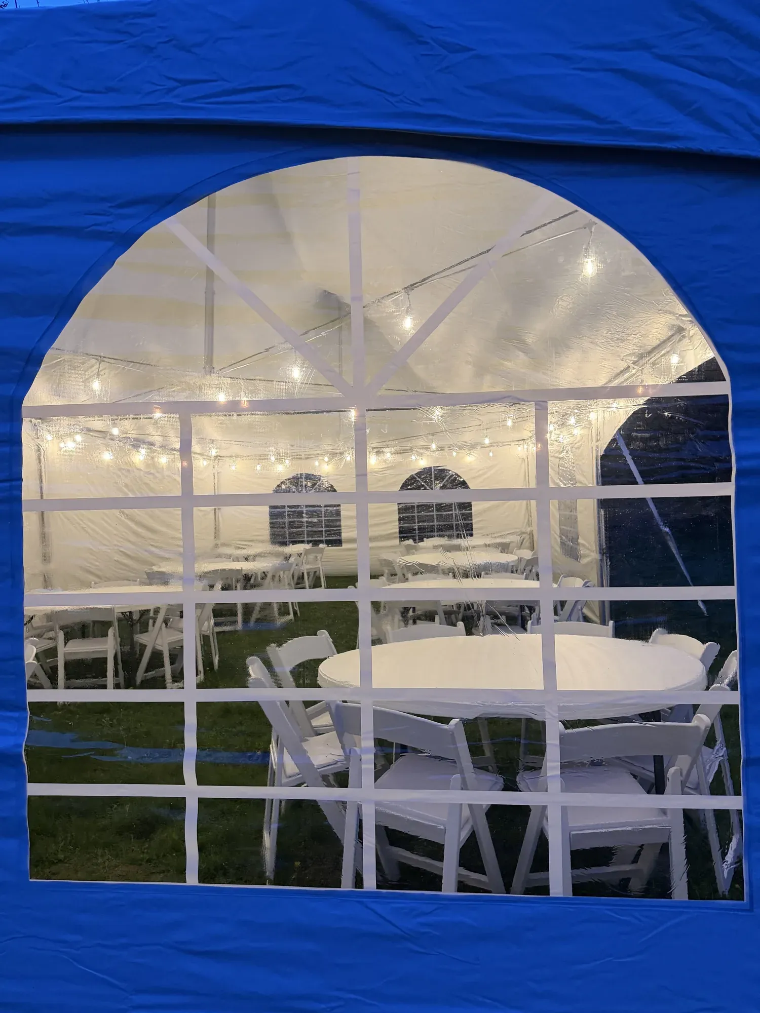 Peeking through the cathedral window of a tent sidewall to see tables and padded resin chairs setup for a wedding.