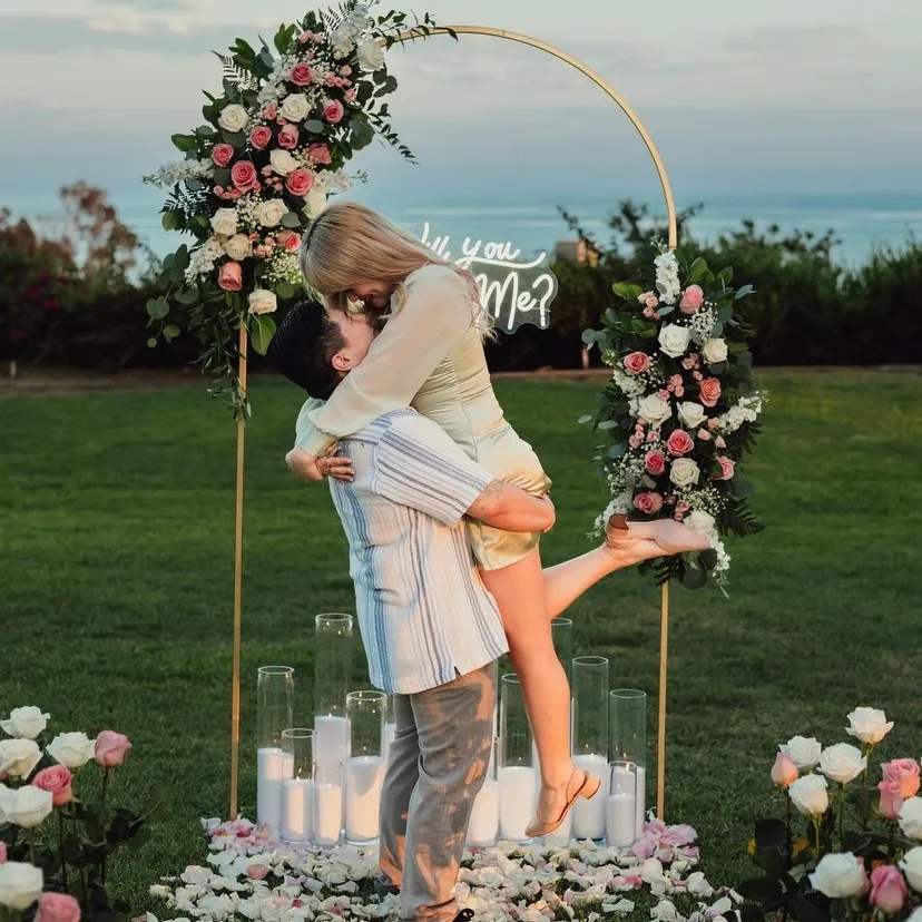 Montaje inspiracional con arco floral de seda, rótulo de neón 'Will You Marry Me?' y iluminación romántica para propuesta de matrimonio.