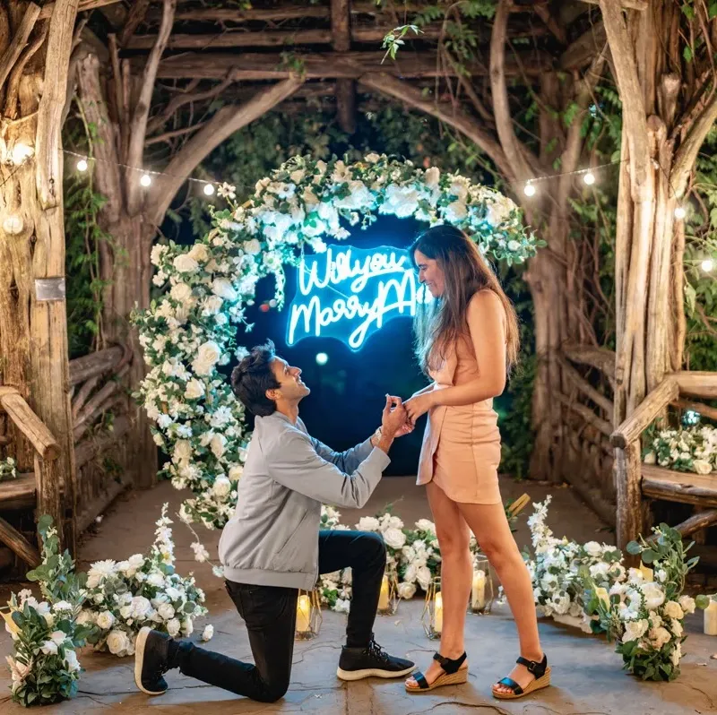 Montaje inspiracional con arco floral de seda, rótulo de neón 'Will You Marry Me?' y iluminación romántica para propuesta de matrimonio.