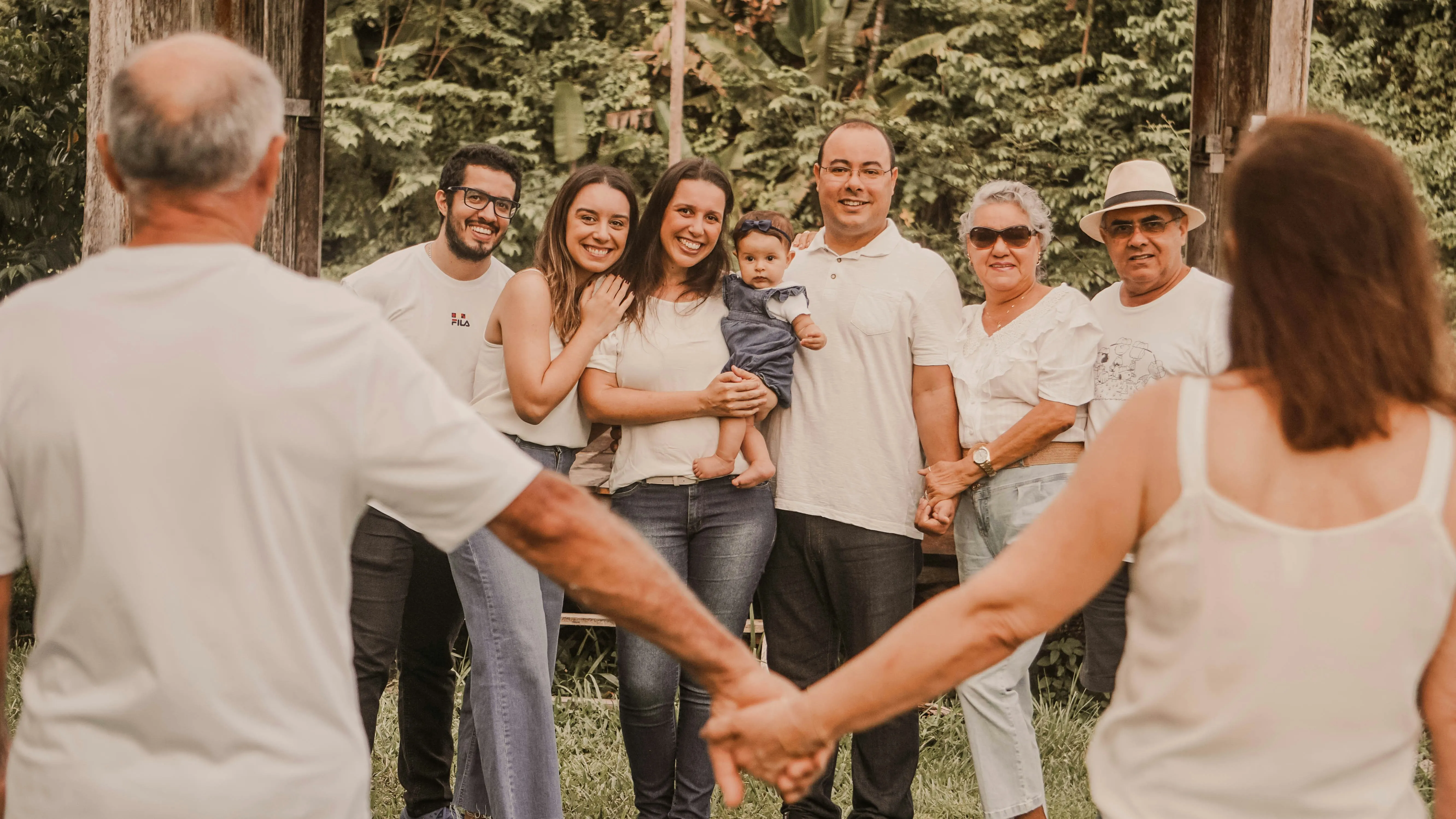 A multi-generational family enjoys an outdoor reunion in a park while playing cornhole, battle buckets, and soccer granny pants. Children, parents, and grandparents laugh together under shade trees with picnic tables and reunion decorations in the background.