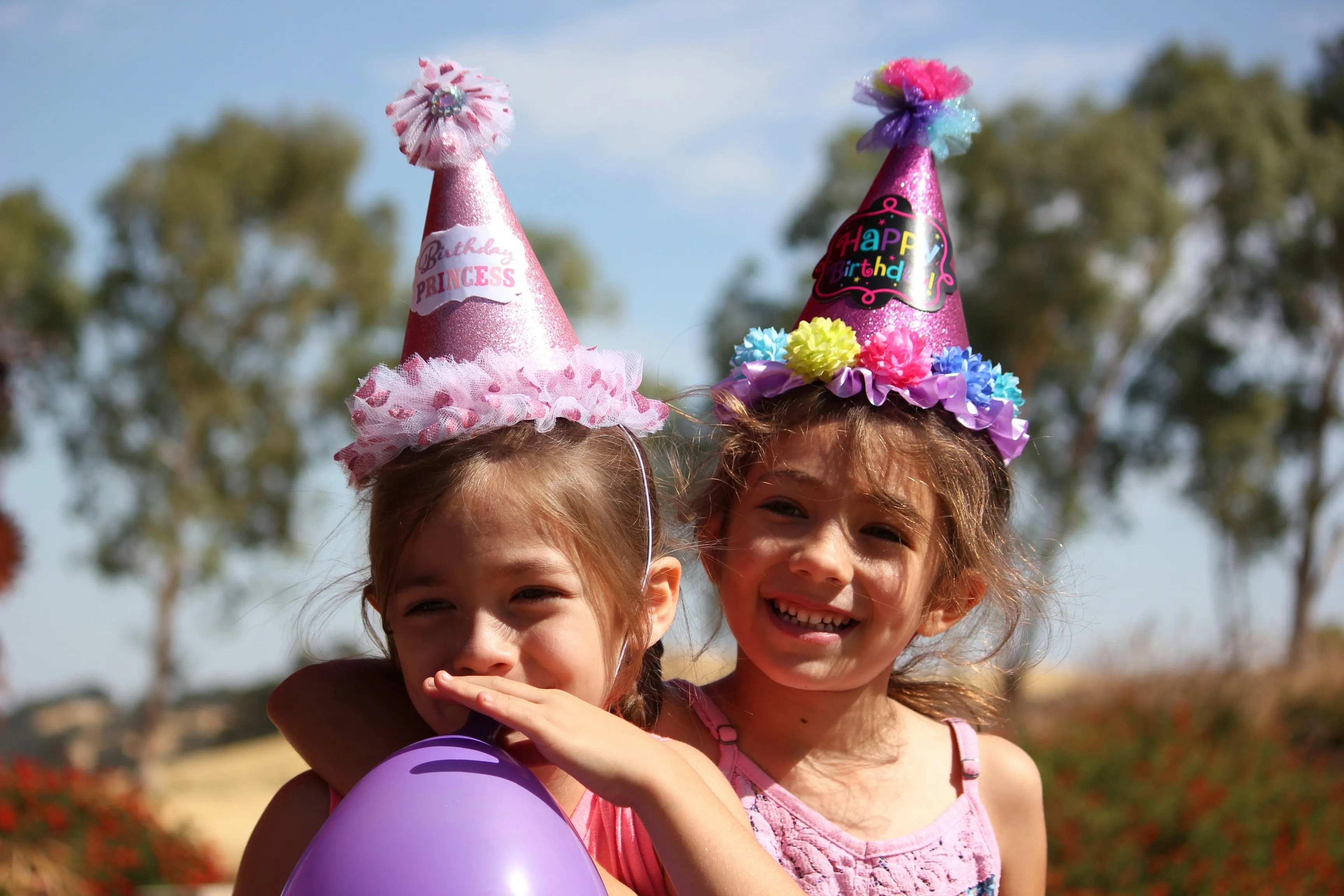 Two young girls wearing colorful birthday party hats smile and celebrate outdoors at a children's birthday party with a purple balloon on a sunny day.