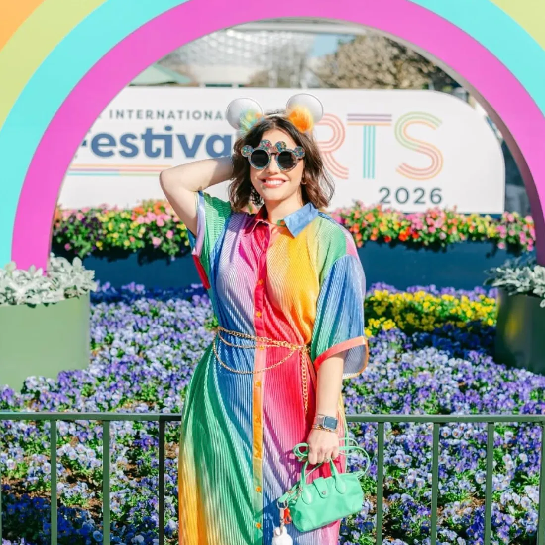 Woman wearing Mickey ear sunglasses and Disney ears headband at EPCOT Festival of the Arts 2026, styled with a rainbow dress, gold chain belt, and green mini bag with bag charm.
