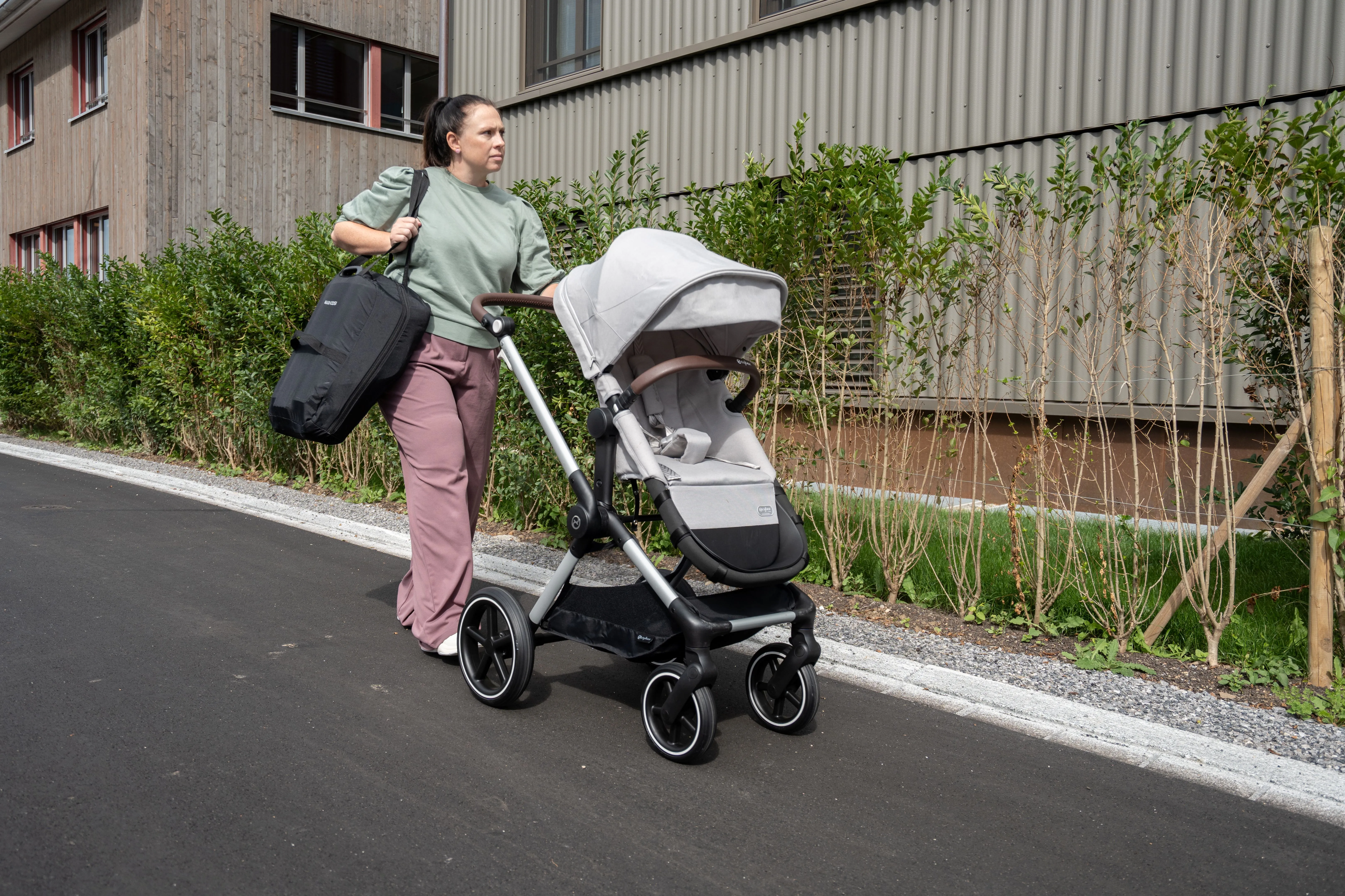 Women with brown hair, green jumper and mauve pants walking down a street. She is pushing a stroller and carrying a car seat in a carry bag over her shoulder.