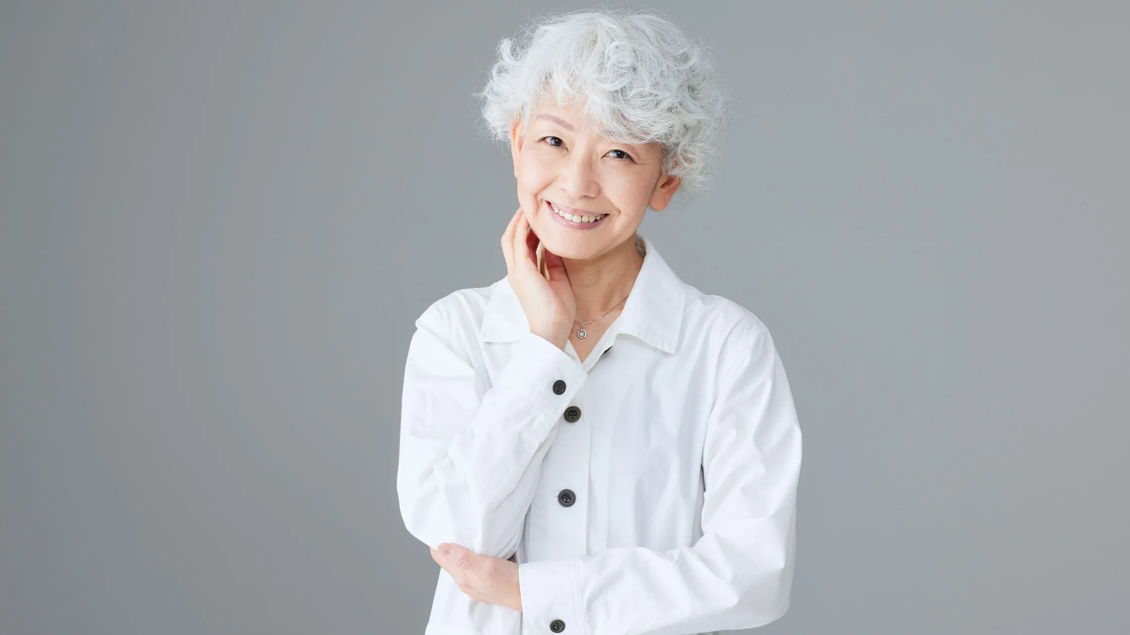A happy woman standing facing the camera, smiling, wearing a long-sleeved, button-down white shirt, against a grey background.