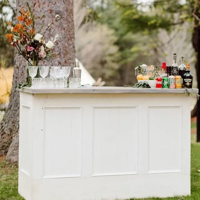 White fold-up bar with trim in a backyard near a tree, stocked with assorted glasses, alcohol bottles, mixers, and fresh fruit for serving drinks.