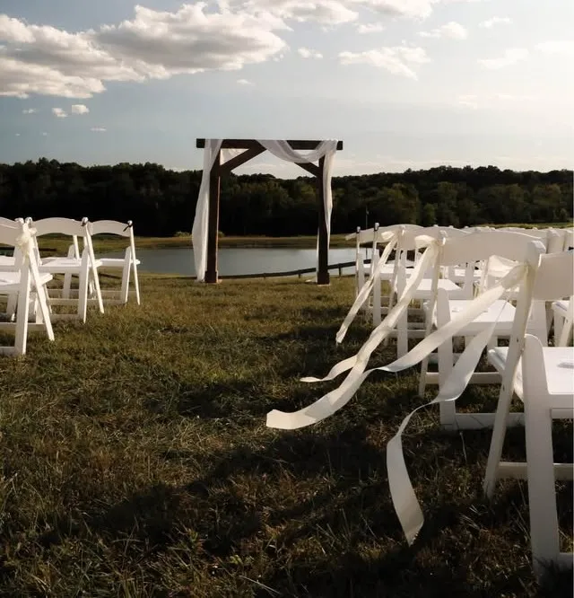 Outdoor wedding aisle with white garden chairs and a cedar arbor decorated with white draping.