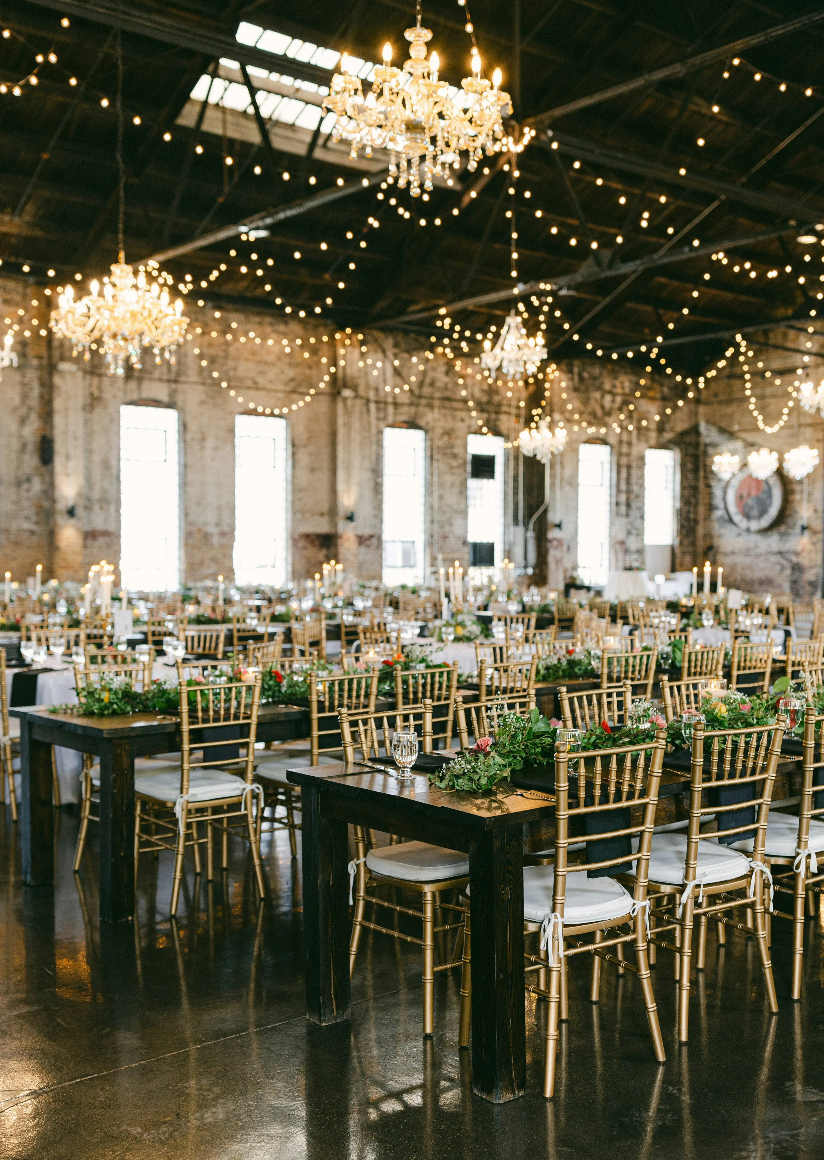 A reception venue set up with gold chairs, greenery on the tables, and market lights and chandeliers hanging from the ceiling.