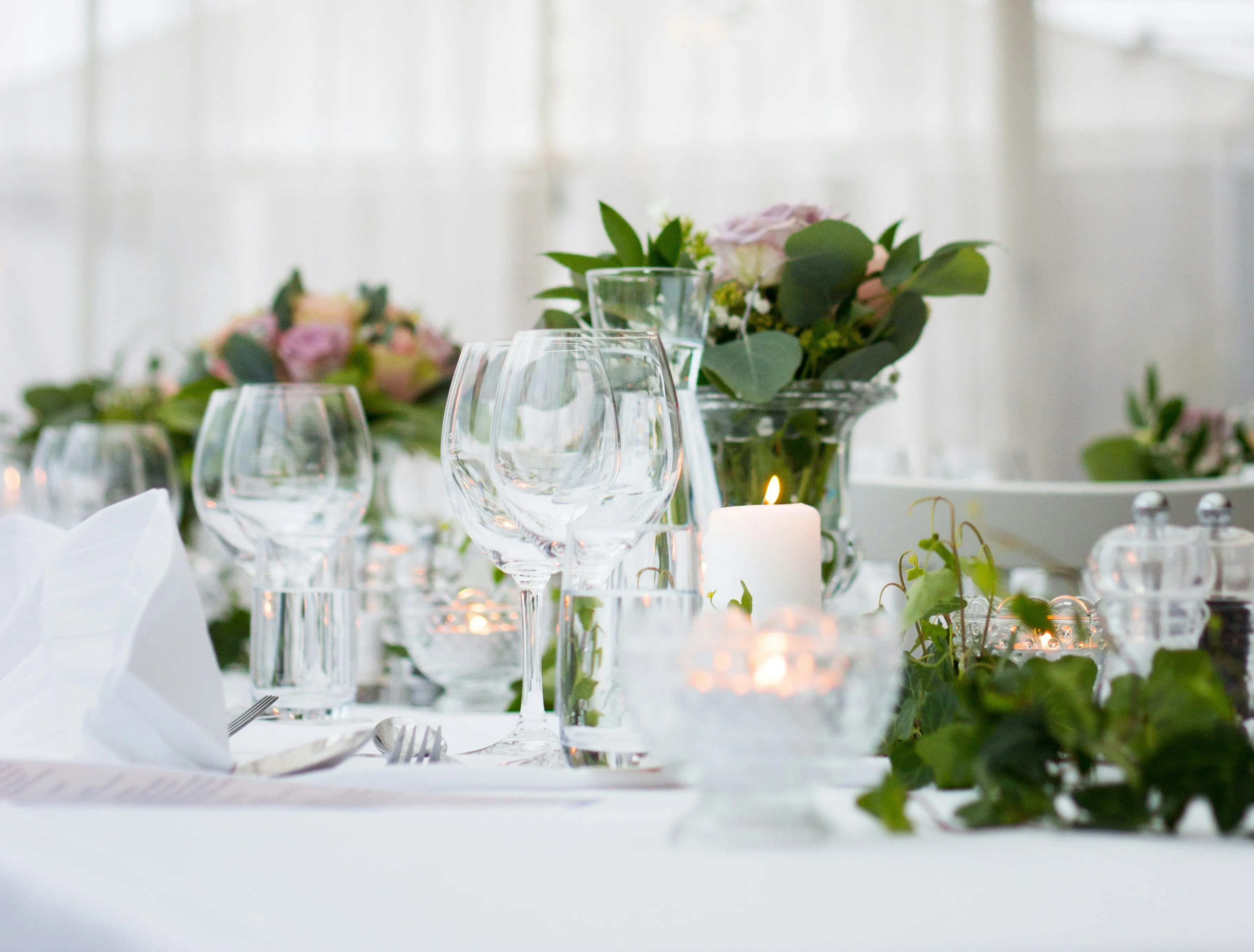 Multiple table settings with wine glasses, candles, utensils, napkins, and floral arrangements.