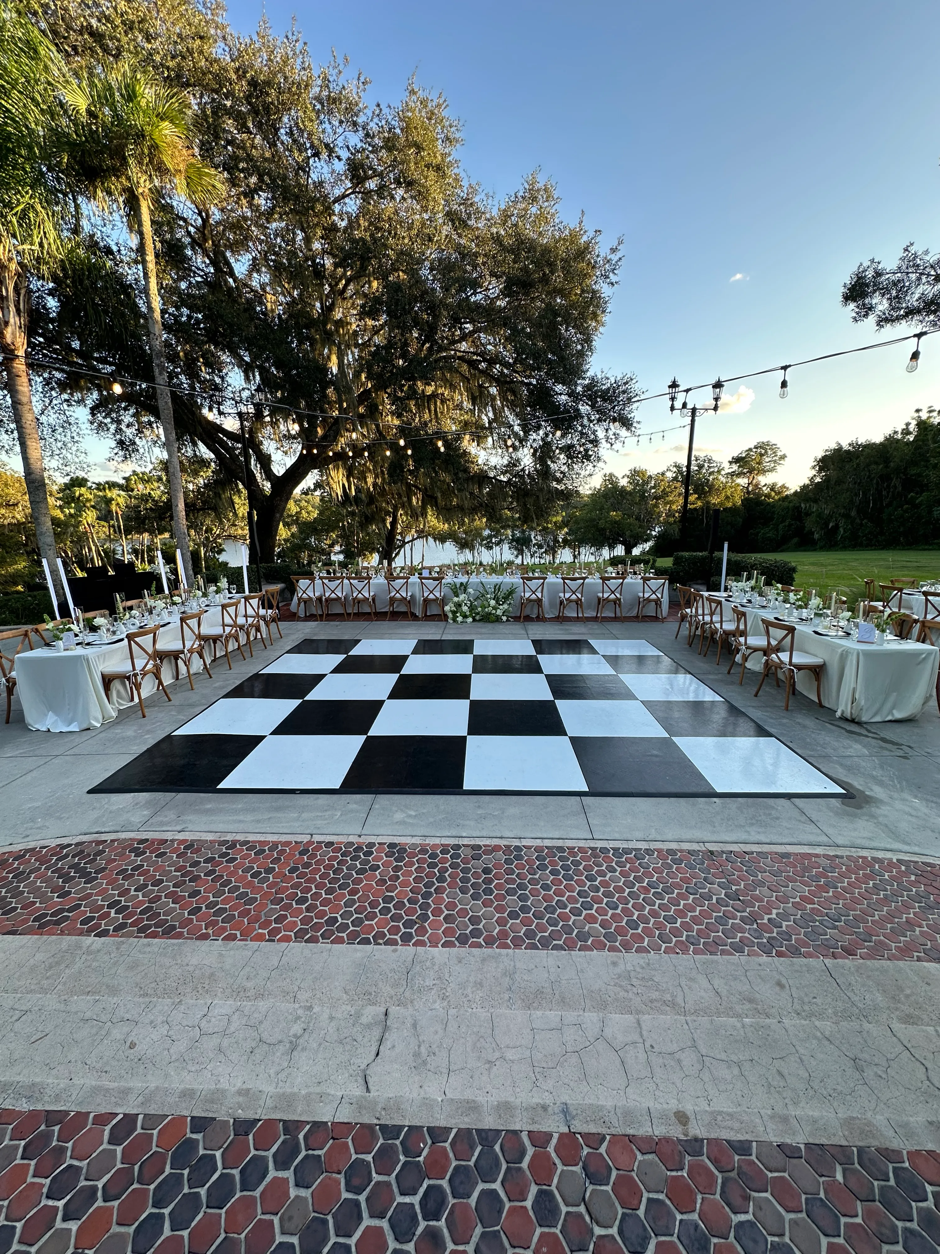 An outdoor reception venue with banquet style tables and chairs surrounding a black and white checkered dance floor.