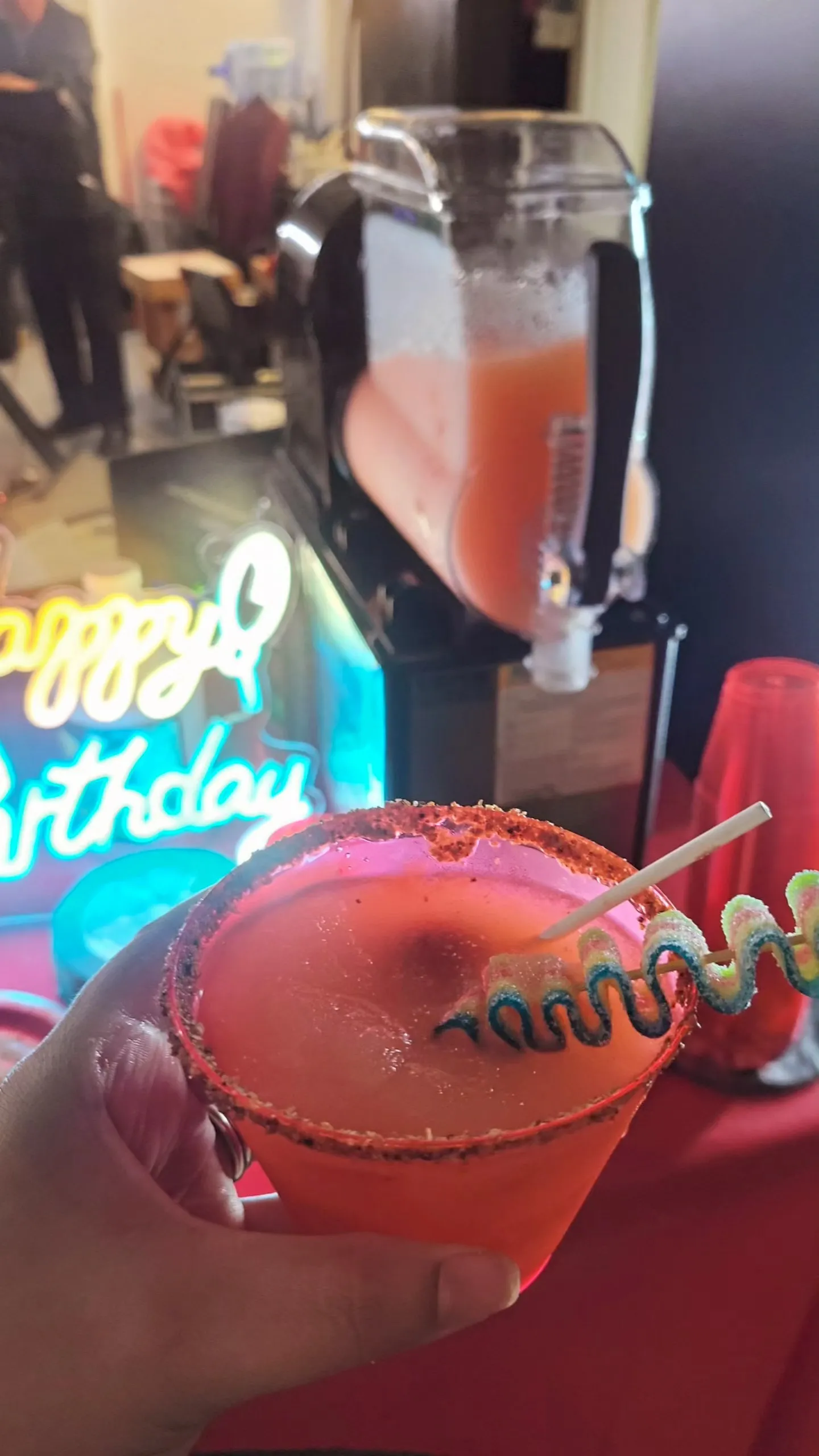 a small female hand holding a cup of slushie with a tajin rim and decorative straw with neon sign and margarita machine in the background