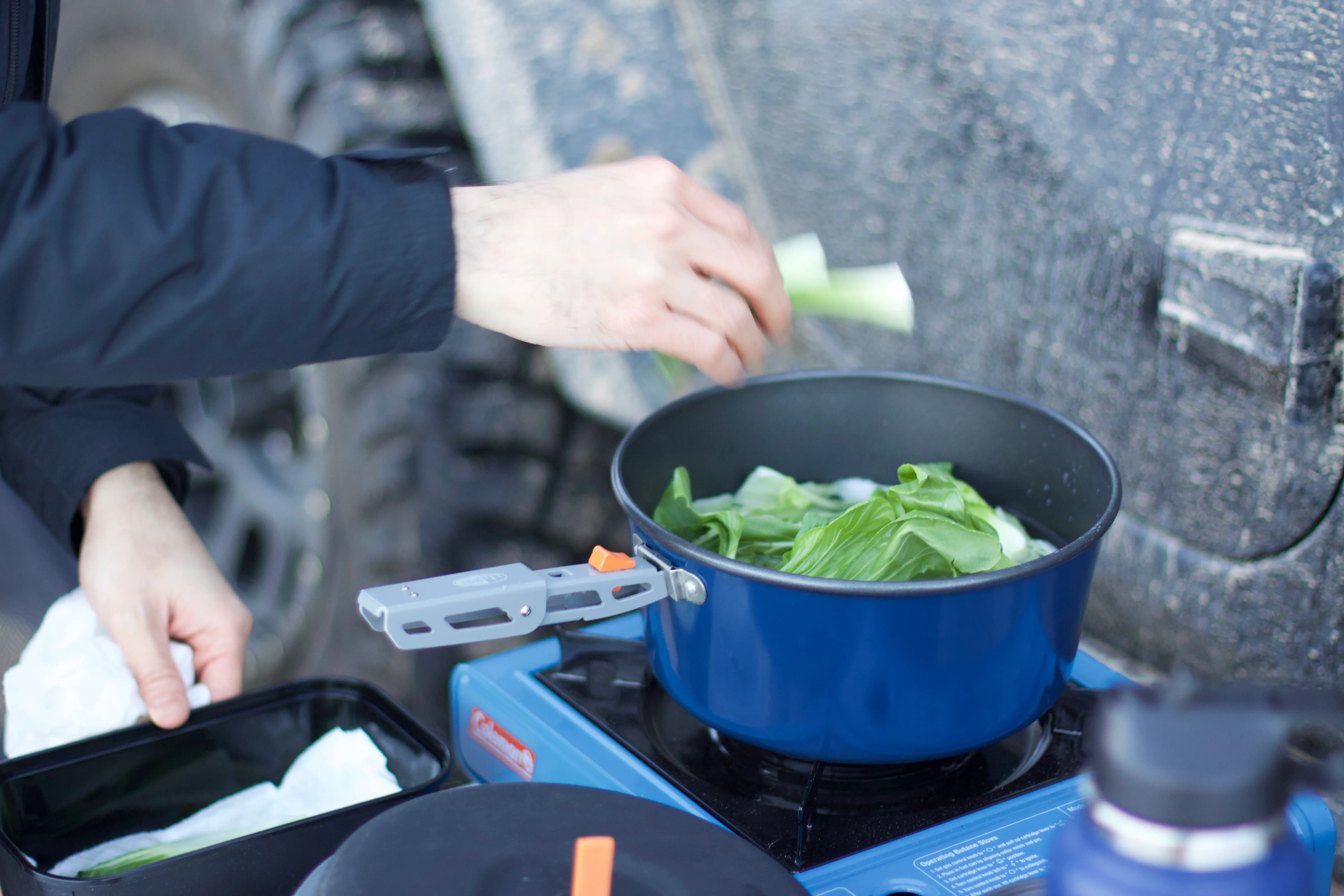Camper cooking vegetables in a pot on a portable camp stove outdoors.