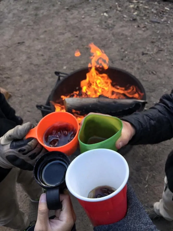 Campers enjoying hot drinks together around a campfire during a weekend camping trip.