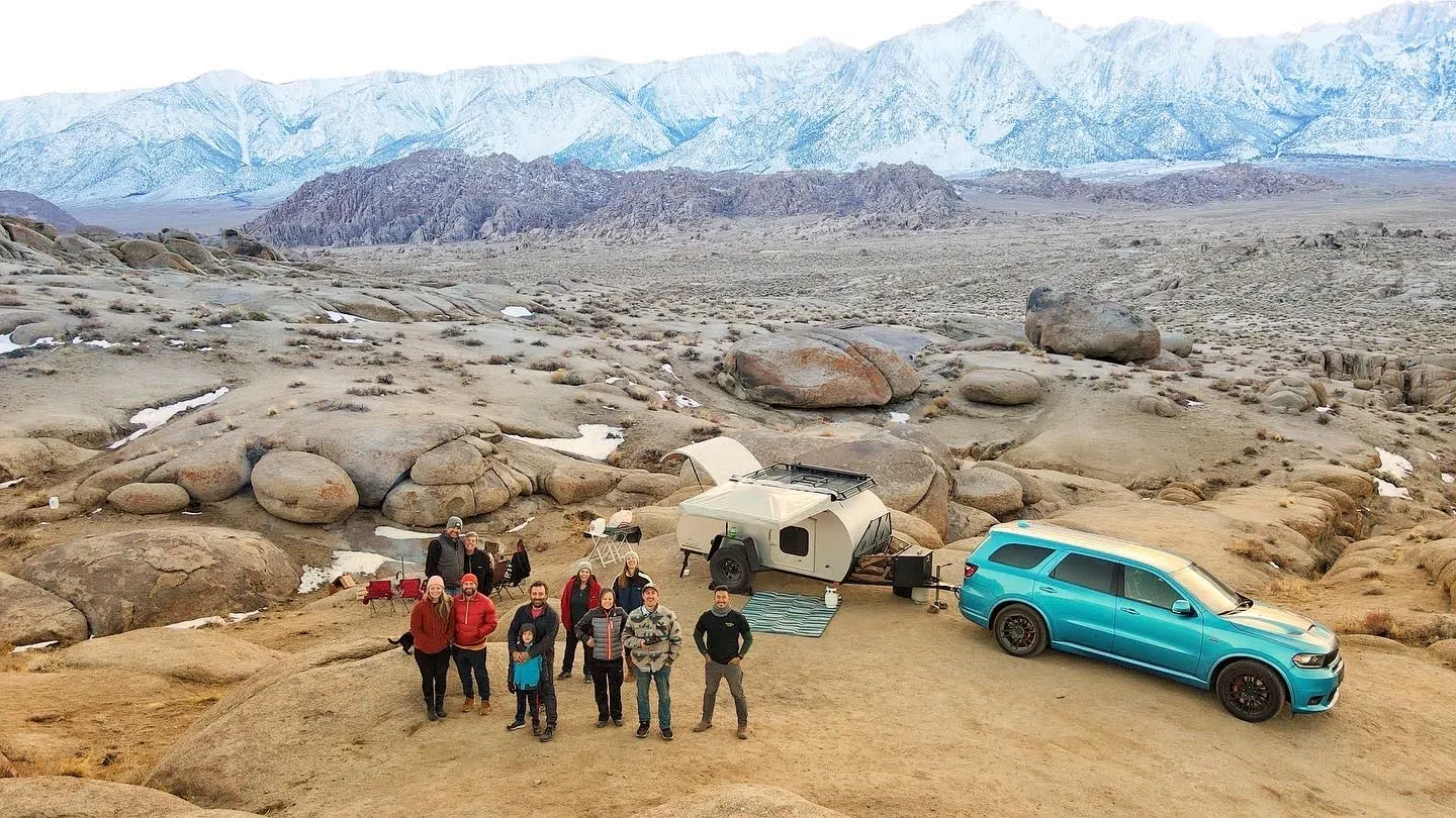 Group of campers standing beside a travel trailer and SUV in a rocky mountain landscape with snow-covered peaks in the distance.
