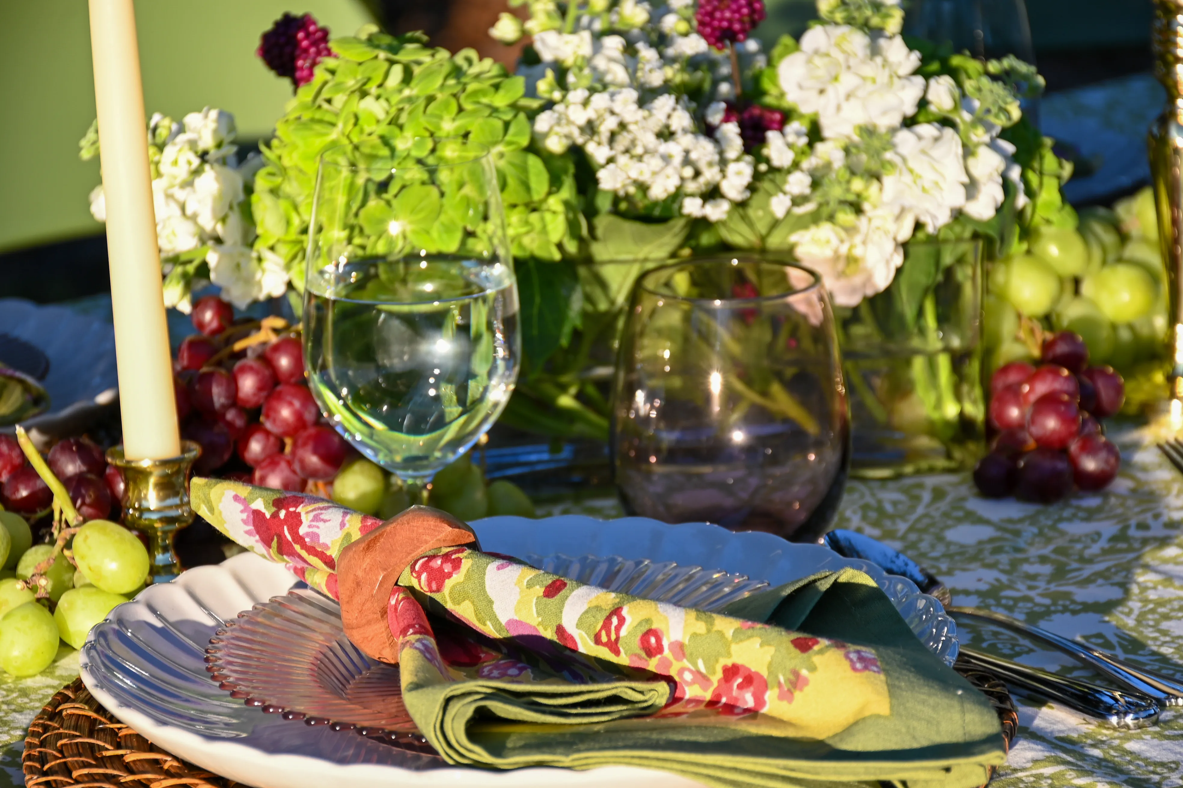 Close-up of an elegant outdoor table setting with green patterned tablecloth, floral napkins, white plates on woven chargers, taper candles, fresh grapes, and garden flowers for a spring or summer tablescape.