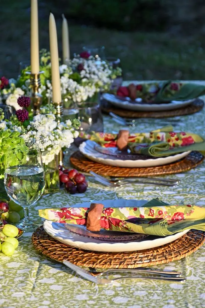 Close-up of an elegant outdoor table setting with green patterned tablecloth, floral napkins, white plates on woven chargers, taper candles, fresh grapes, and garden flowers for a spring or summer tablescape.