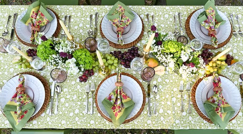 Elegant outdoor dining table set with green floral tablecloth, coordinated napkins, white plates, candles, and fresh grapes, styled for a garden party or spring tablescape.