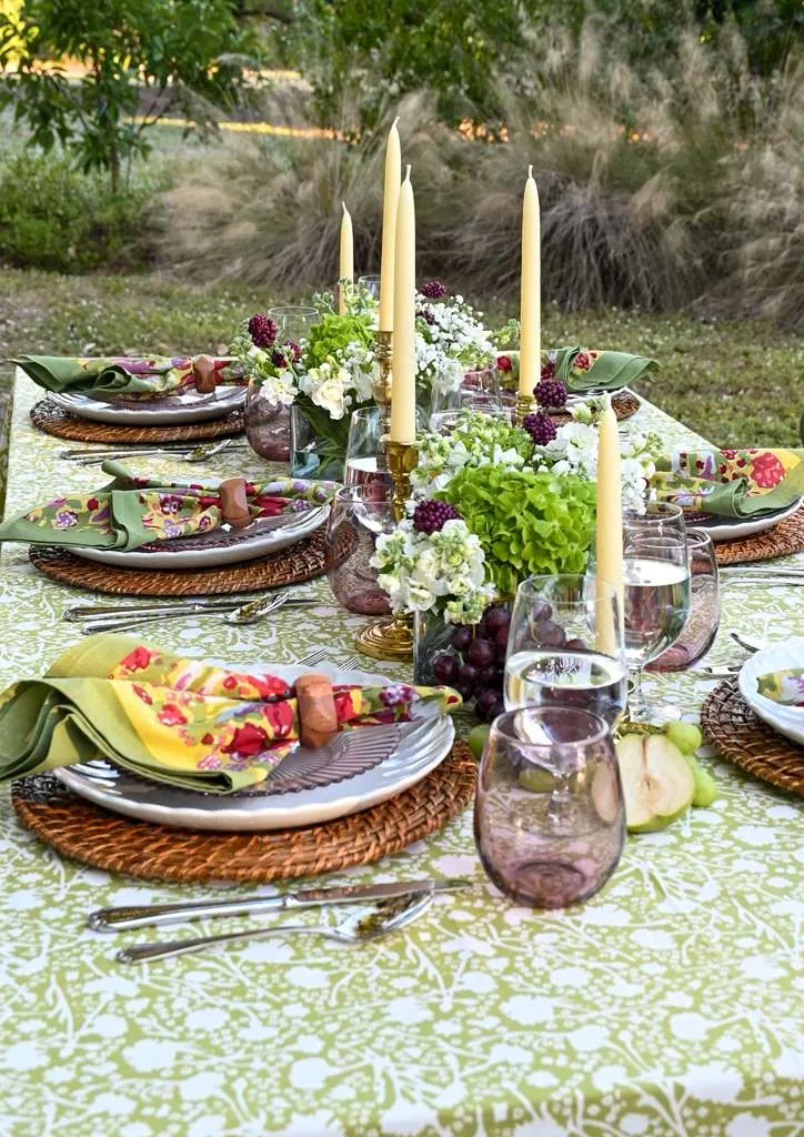 Elegant outdoor dining table set with green floral tablecloth, coordinated napkins, white plates, candles, and fresh grapes, styled for a garden party or spring tablescape.