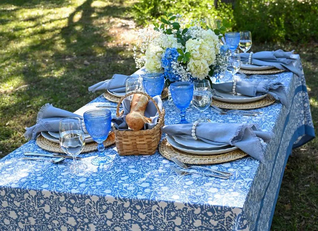Outdoor dining table styled with blue patterned tablecloth, woven chargers, soft gray napkins, blue glassware, fresh bread basket, and white and blue floral centerpiece, set for an elegant garden lunch or summer tablescape.