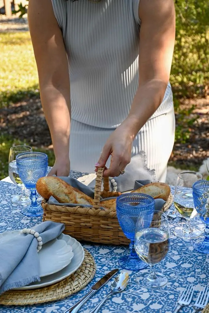 Women placing a bread basket on an outdoor dining table styled with blue patterned tablecloth, woven chargers, soft gray napkins, blue glassware, and white and blue floral centerpiece, set for an elegant garden lunch or summer tablescape.