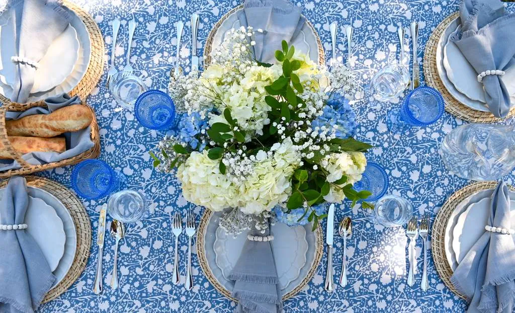 Outdoor dining table styled with blue patterned tablecloth, woven chargers, soft gray napkins, blue glassware, fresh bread basket, and white and blue floral centerpiece, set for an elegant garden lunch or summer tablescape.