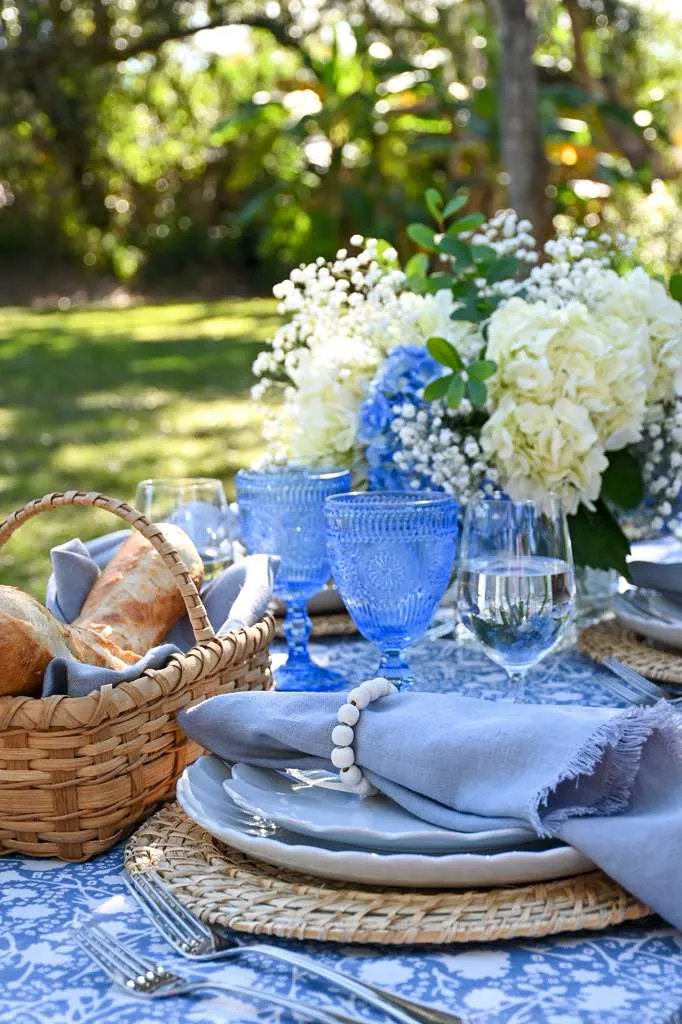 Close up of outdoor dining table styled with blue patterned tablecloth, woven chargers, soft gray napkins, blue glassware, fresh bread basket, and white and blue floral centerpiece, set for an elegant garden lunch or summer tablescape.