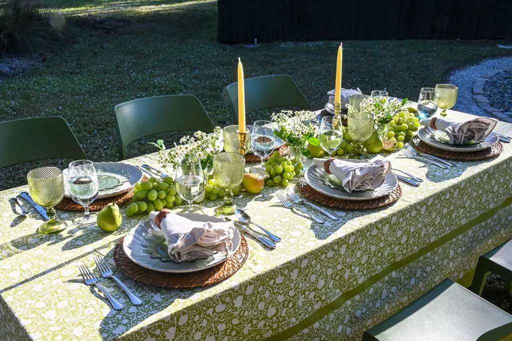 Elegant outdoor garden dining table with green patterned tablecloth, coordinated green chairs, woven chargers, candles, fresh fruit accents, and soft gray napkins styled for a spring or summer alfresco tablescape.