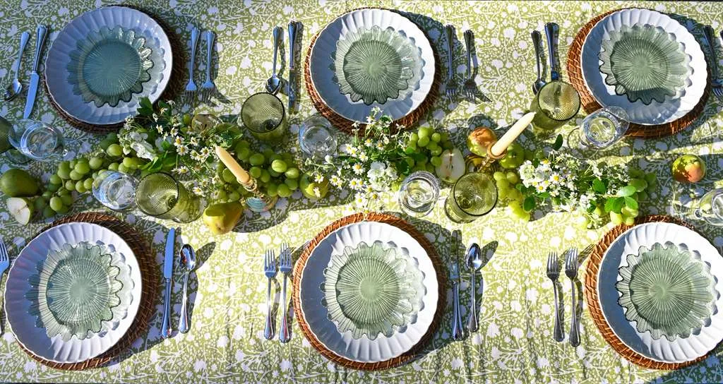 Elegant outdoor garden dining table with green patterned tablecloth, coordinated green chairs, woven chargers, candles, fresh fruit accents, and soft gray napkins styled for a spring or summer alfresco tablescape.