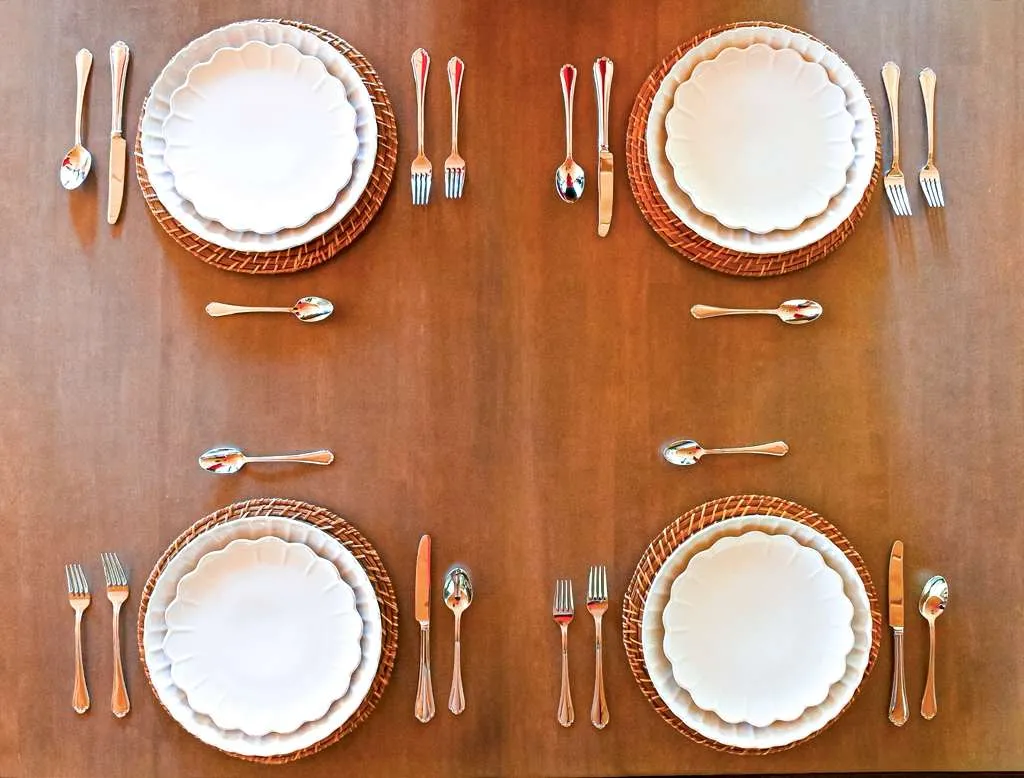 Overhead view of four elegant place settings with scalloped white plates on woven chargers and polished silver flatware arranged on a warm wood dining table for a classic, minimalist tablescape.