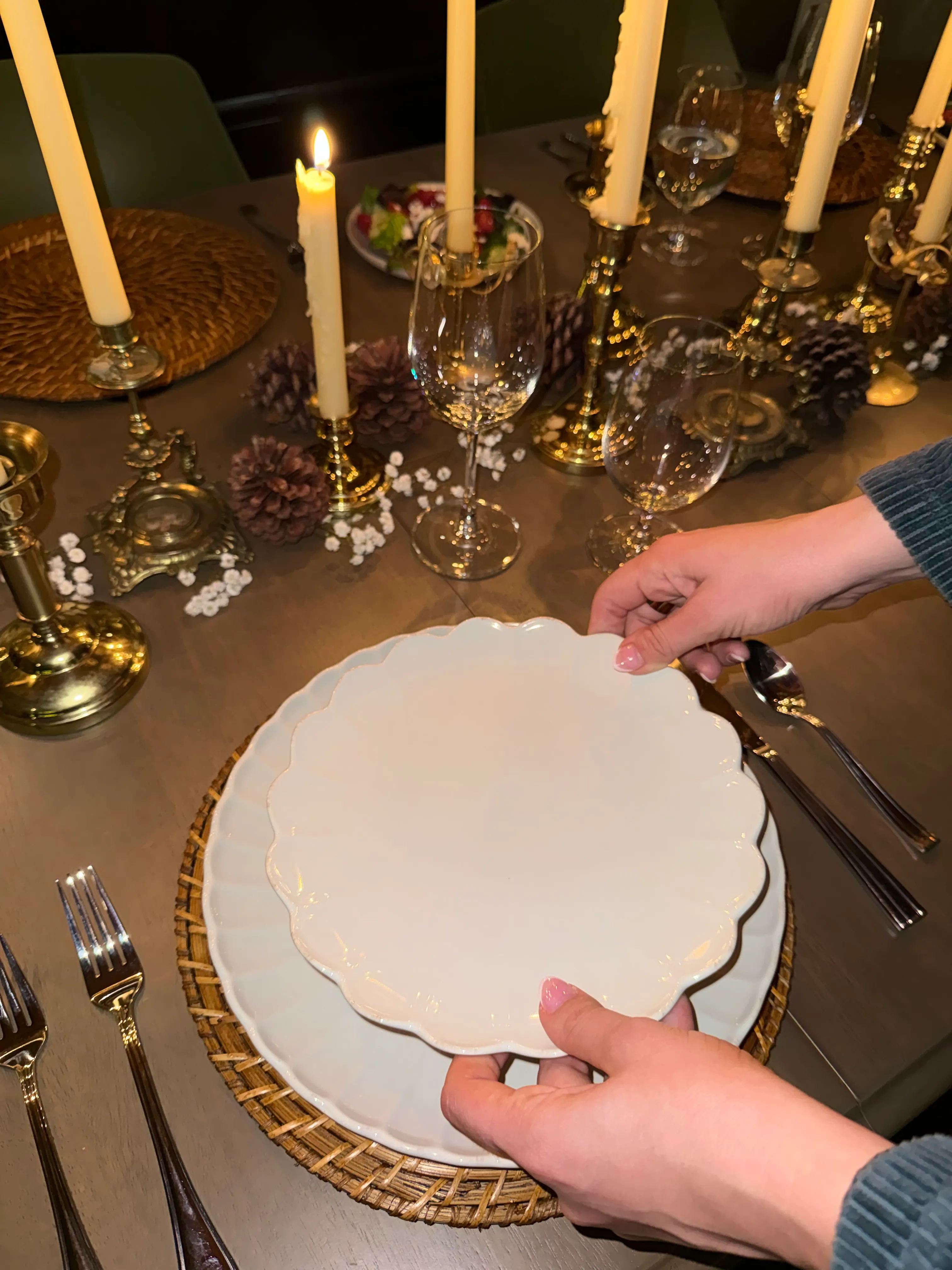 Person arranging an elegant dinner place setting with scalloped white plates on woven charger, candlelit table with gold candlesticks, wine glasses, pinecone accents, and warm seasonal tablescape decor.