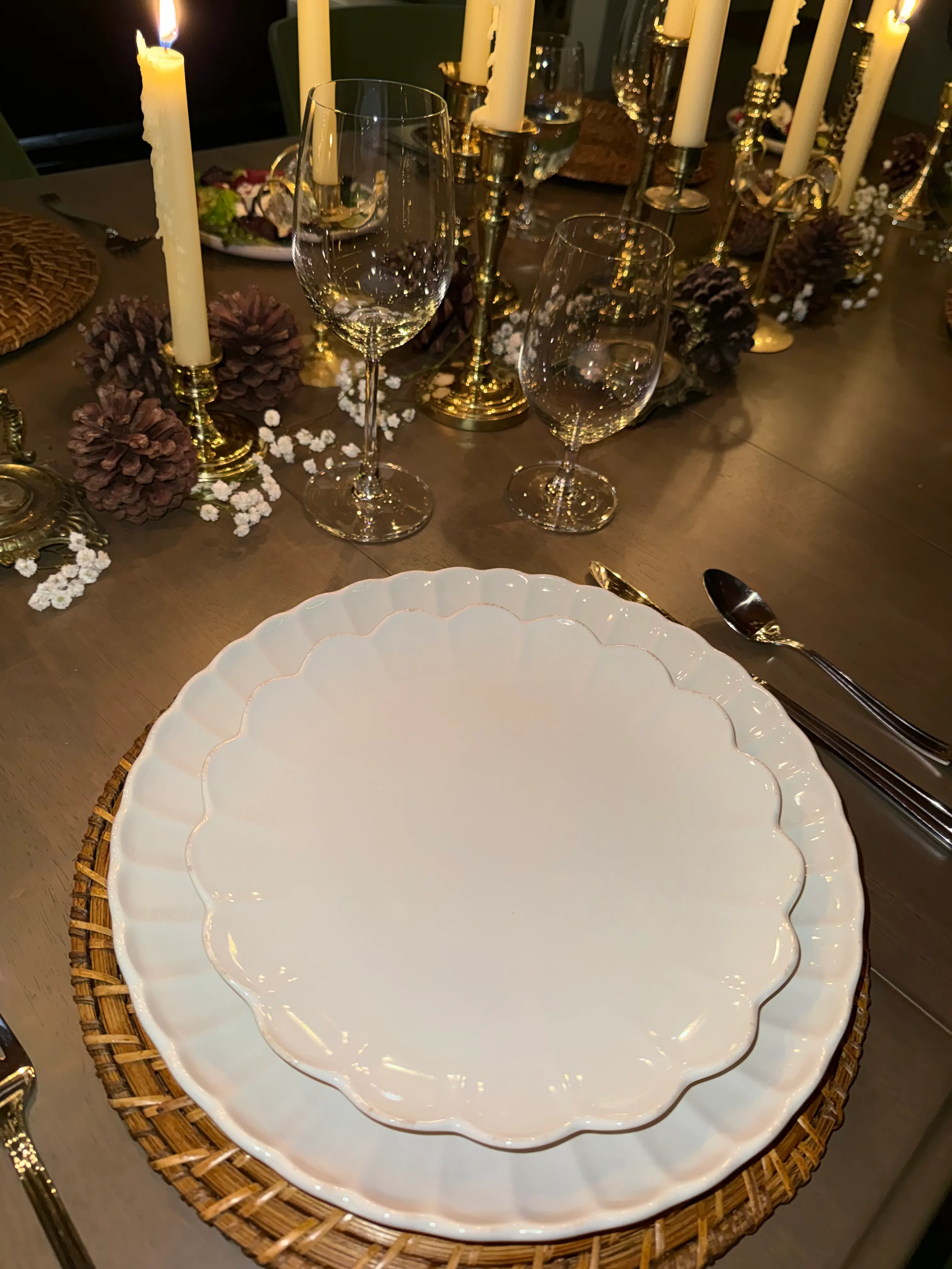 Elegant fall or holiday dinner place setting with scalloped white plates on woven charger, candlelit table with gold candlesticks, wine glasses, pinecone accents, and warm seasonal tablescape decor.