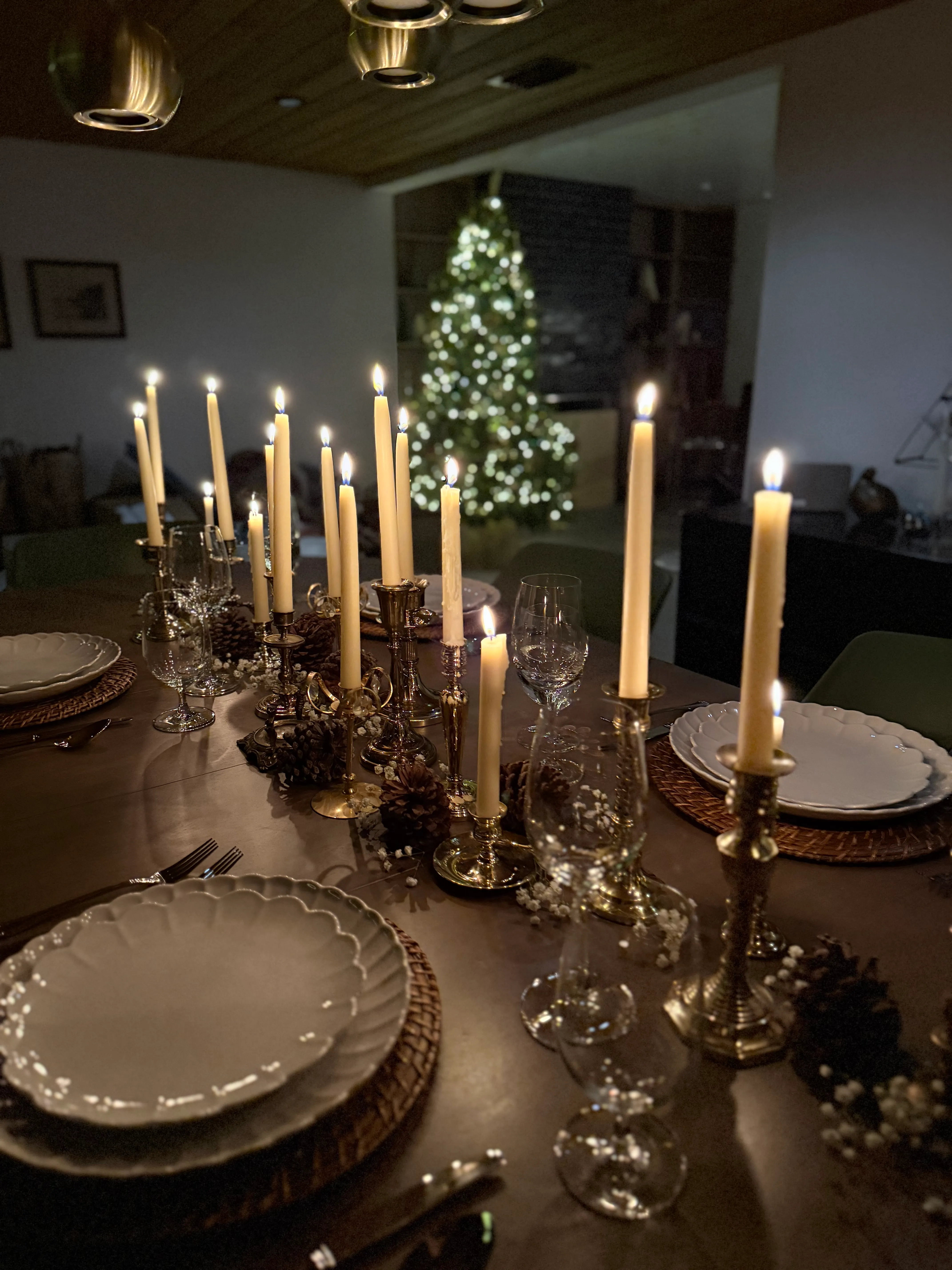 Elegant fall or holiday dinner place setting with scalloped white plates on woven charger, candlelit table with gold candlesticks, wine glasses, pinecone accents, and warm seasonal tablescape decor.