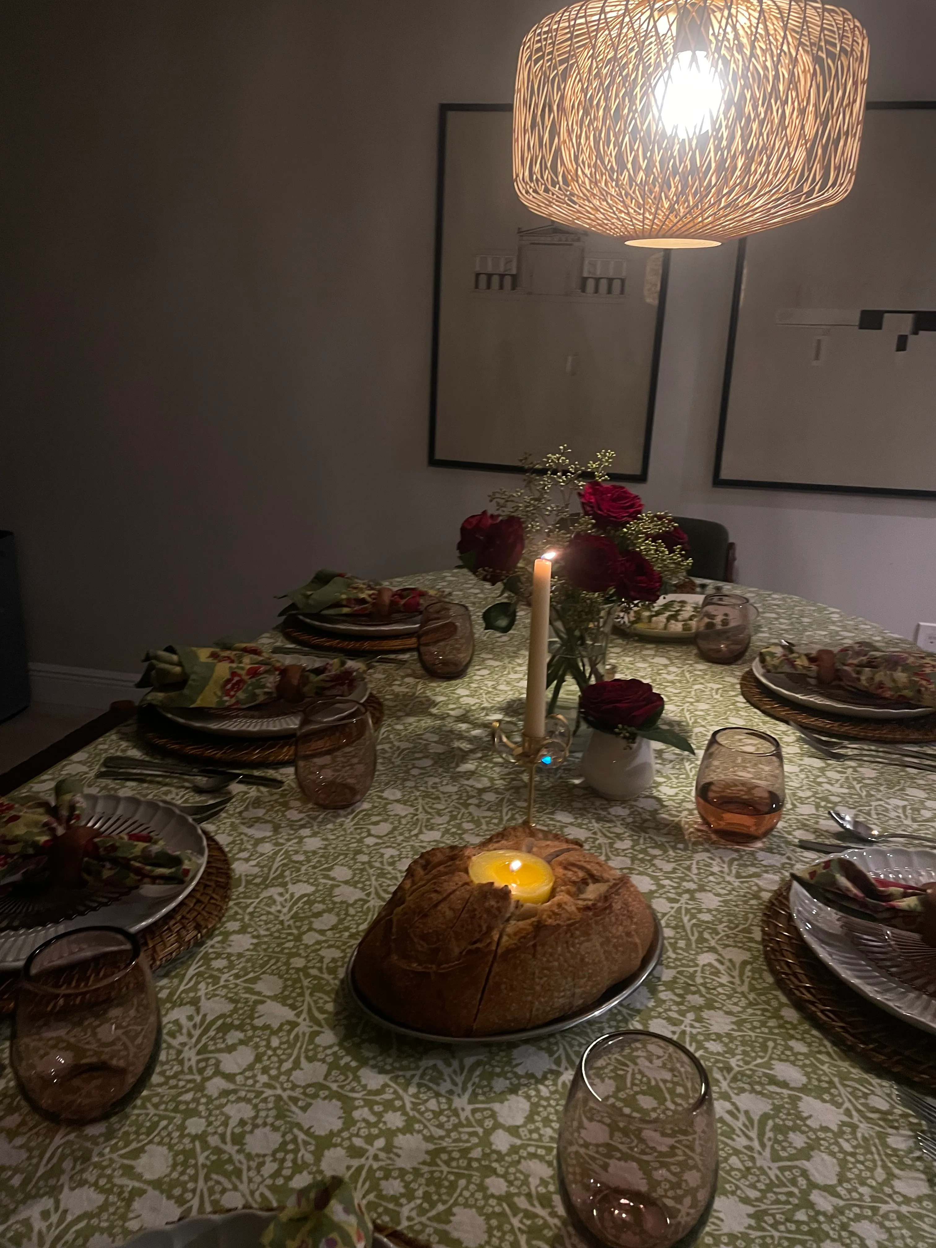 Cozy indoor dining table set with green patterned tablecloth, woven chargers, floral napkins, candlelight centerpiece with red roses, artisan bread, and warm pendant lighting for an intimate dinner tablescape.