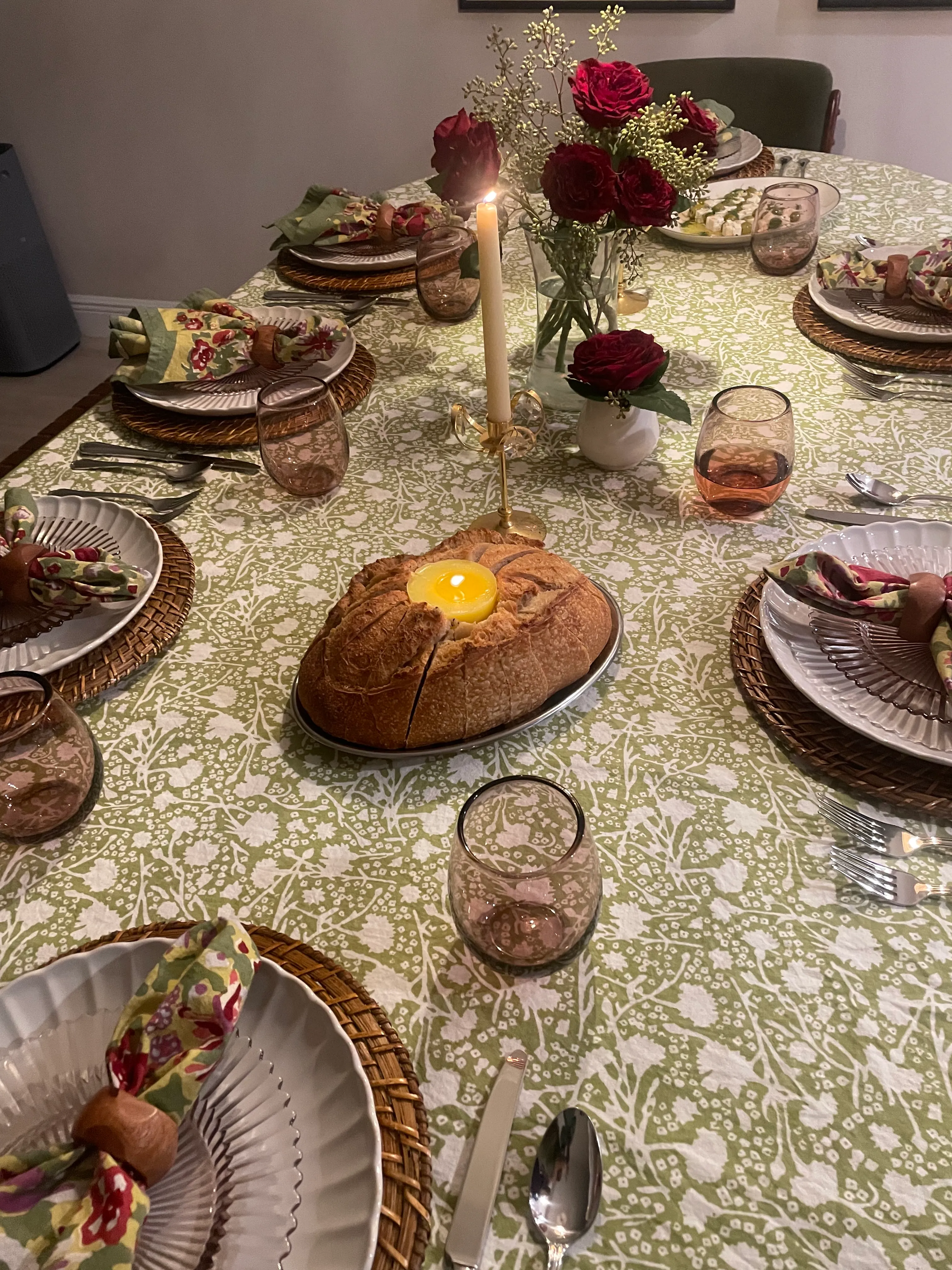 Cozy indoor dining table set with green patterned tablecloth, woven chargers, floral napkins, candlelight centerpiece with red roses, artisan bread, and warm lighting for an intimate dinner tablescape.