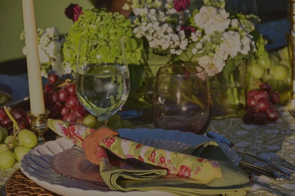 Close-up of an elegant outdoor table setting with green patterned tablecloth, floral napkins, white plates on woven chargers, taper candles, fresh grapes, and garden flowers for a spring or summer tablescape.