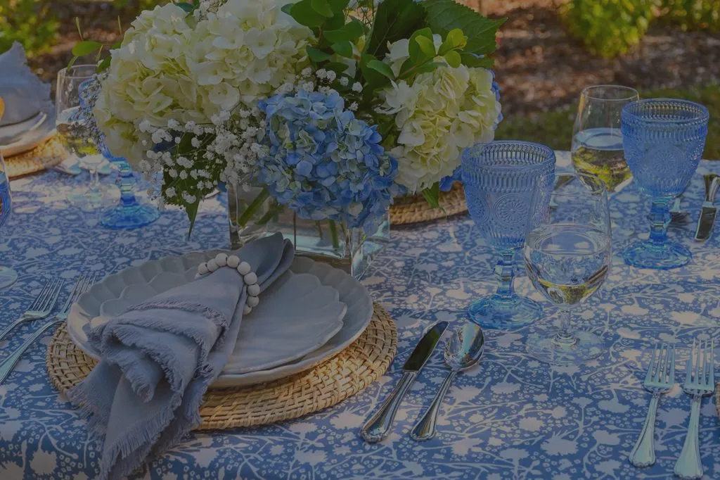 Close up of outdoor dining table styled with blue patterned tablecloth, woven chargers, soft blue-gray napkins, blue glassware, and white and blue floral centerpiece, set for an elegant garden lunch or summer tablescape.
