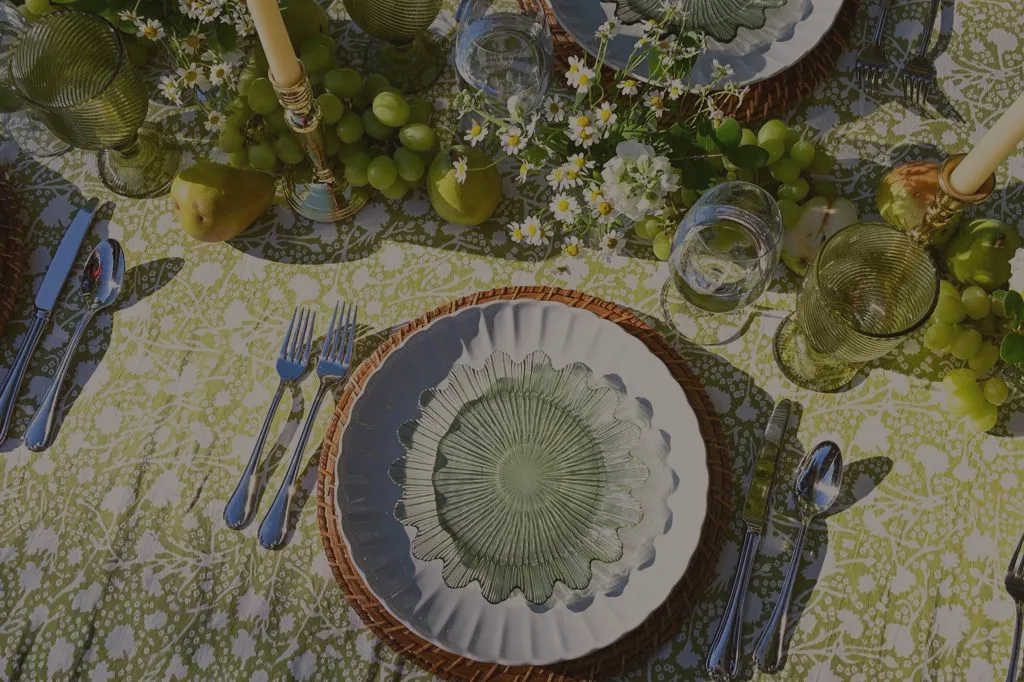 Elegant outdoor garden dining table with green patterned tablecloth, coordinated green chairs, woven chargers, candles, fresh fruit accents, and soft gray napkins styled for a spring or summer alfresco tablescape.