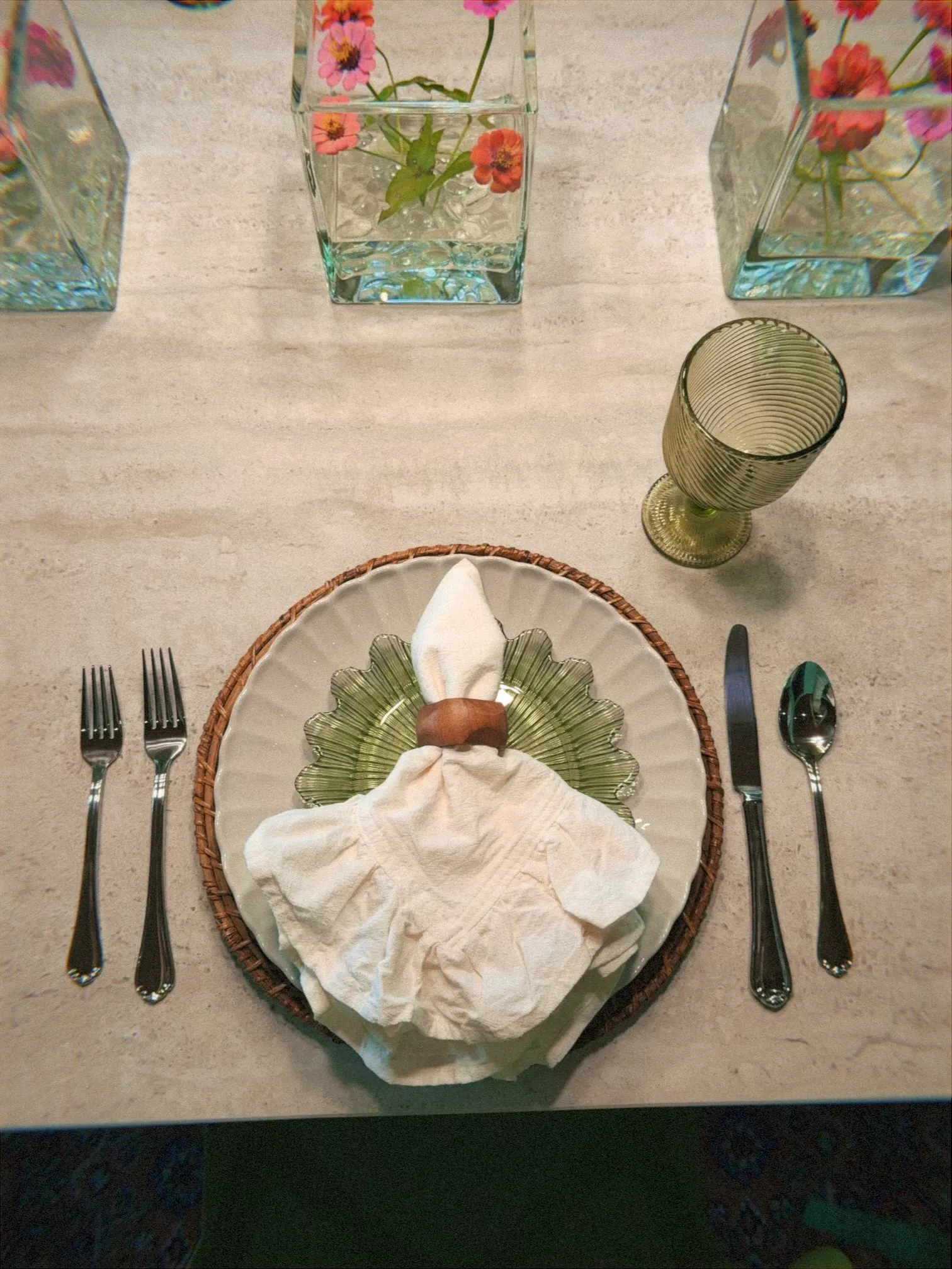 A close-up view of an elegant table setting on a light stone dining table. The place setting features a woven round placemat topped with a large white scalloped dinner plate and a smaller green leaf-shaped glass plate. A white cloth napkin is loosely gathered through a wooden napkin ring and rests in the center. Two forks are placed on the left, with a knife and spoon on the right, alongside a textured green goblet. In the background, square glass vases filled with water and small pink and orange flowers decorate the center of the table.