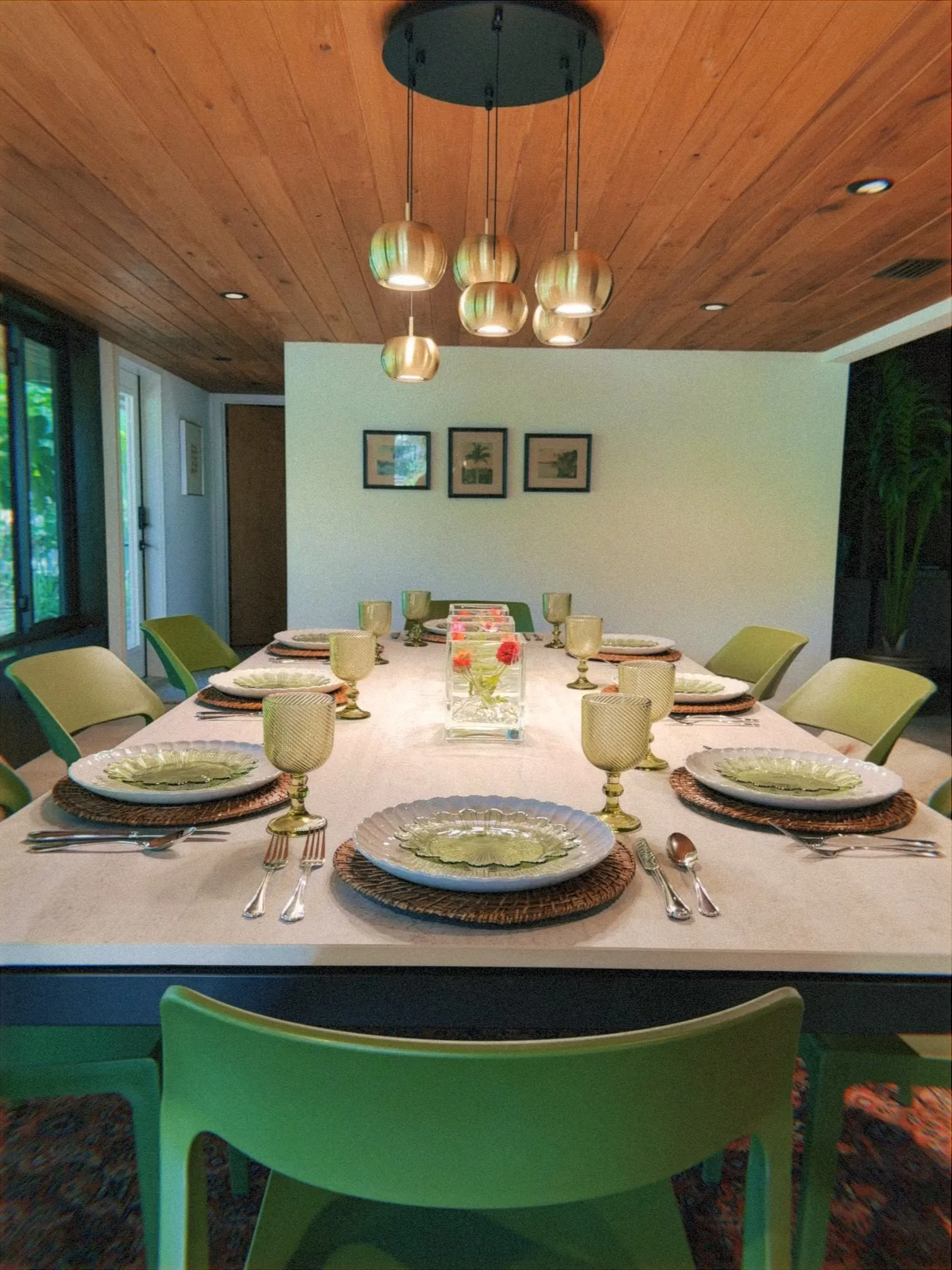 A modern dining room with a long white rectangular table set for eight, featuring green chairs, woven placemats, white scalloped plates, and textured green goblets. A clear glass vase with small red flowers serves as the centerpiece. Above the table hangs a cluster of round pendant lights with warm-toned shades, suspended from a circular black ceiling mount. The room has a wood-paneled ceiling with recessed lighting, framed artwork on the back wall, and large windows along one side that bring in natural light.