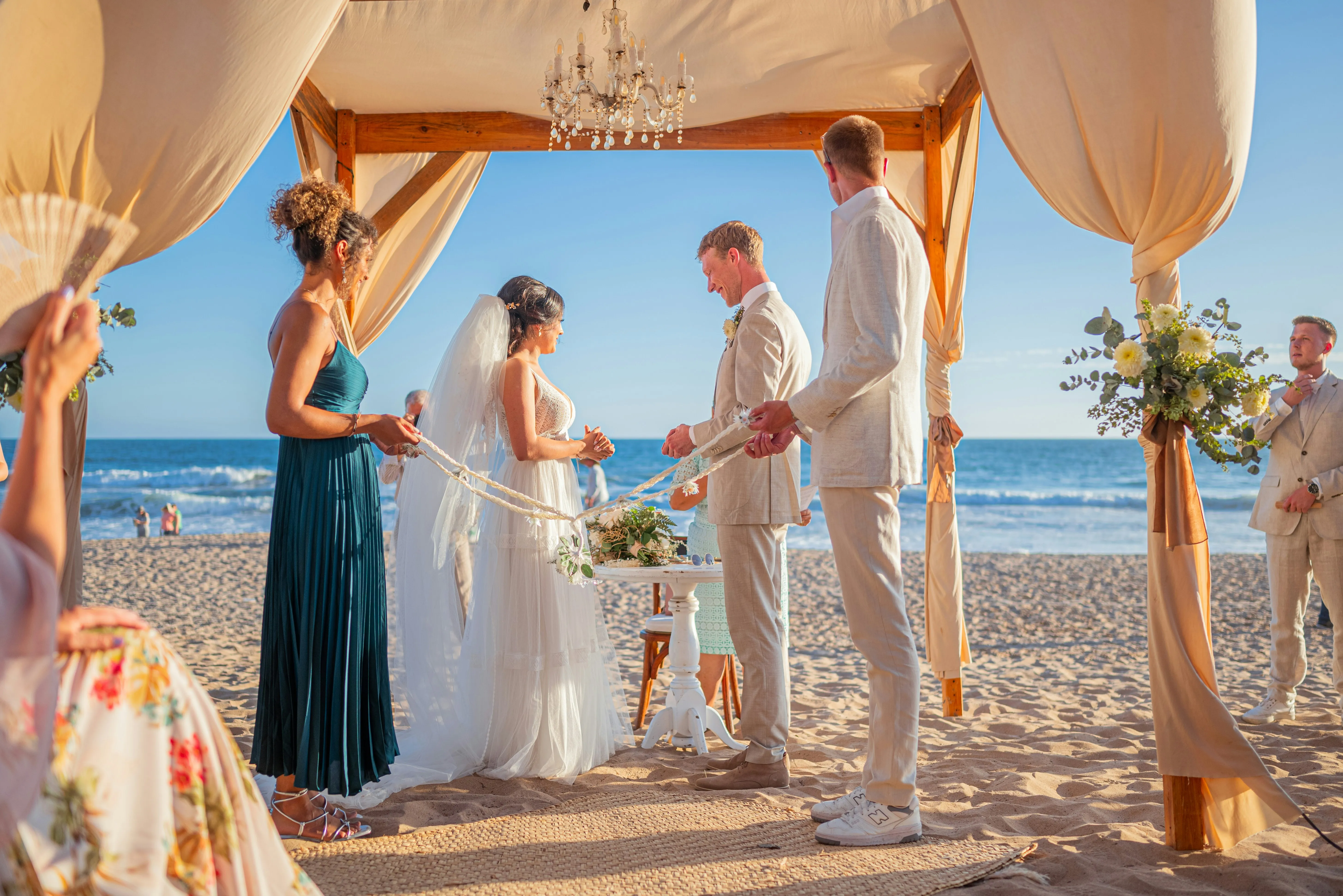 A bride, groom, maid-of-honor, and best man performing a handfasting while standing under a tent at the beach.