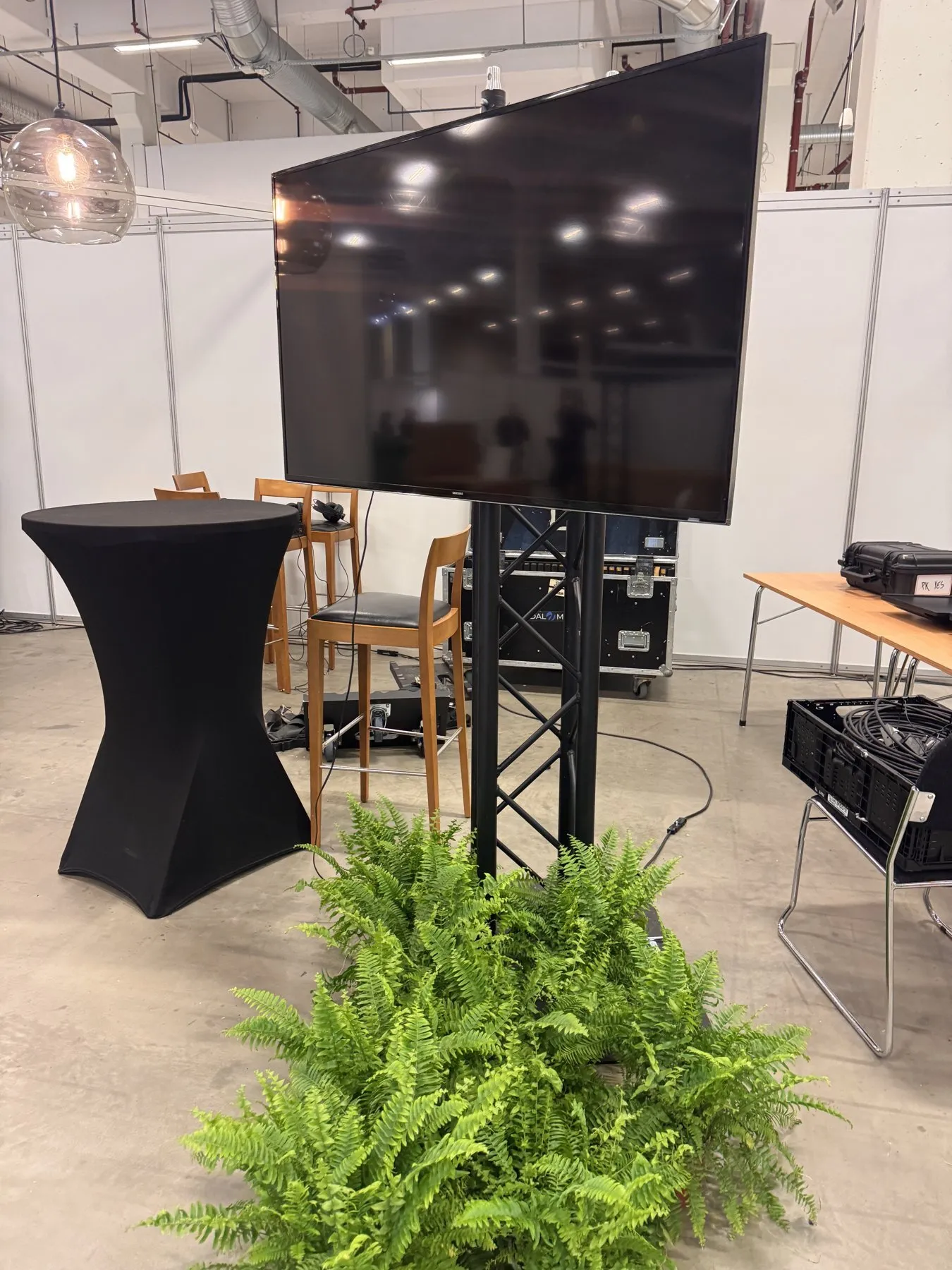 TV-skjerm på truss-stativ med cocktailbord og planter på messestand