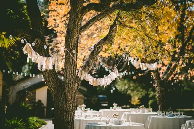 Outdoor event with round banquet tables beneath large trees decorated with string lights and fabric garlands, creating a beautiful backyard party setting.